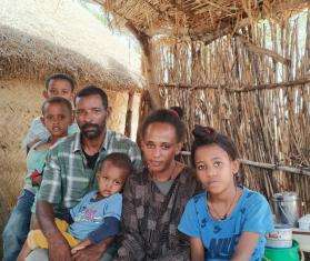 A family of four kids and a mother and father sitting under a thatched roof looking at the camera. 